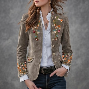 May include: A taupe suede blazer featuring intricate floral embroidery in shades of orange, green, and red along the lapels, cuffs, and pockets. The jacket includes a row of metallic buttons, worn over a white button-down shirt and dark blue jeans.