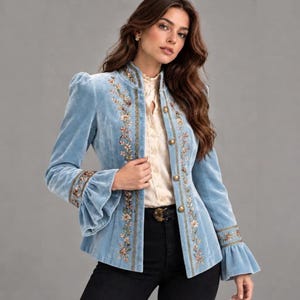 Puede incluir: Una chaqueta de terciopelo azul claro con cuello alto, mangas largas con puños acampanados y bordados florales verticales en los paneles frontales. La chaqueta se cierra con una hilera de botones dorados y se lleva sobre una camisa color crema.