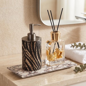 Puede incluir: Un dispensador de jabón cilíndrico con un patrón de cebra en negro y dorado y una bomba de metal cepillado, junto a un difusor de varillas de vidrio con copos dorados y varillas negras, sobre una bandeja rectangular de mármol blanco y gris.