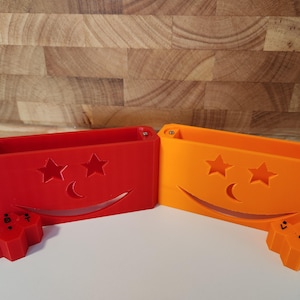 May include: A red and an orange rectangular plastic container, each featuring a cutout design of two stars and a crescent moon above a curved smile. Each container is accompanied by a small, matching hexagonal piece marked with the letters V, B, and T.