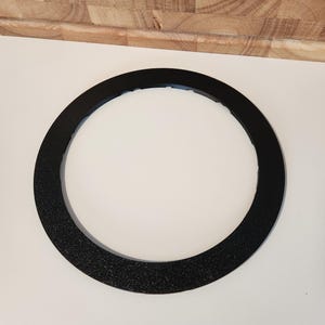 May include: A circular black ring with a textured, matte finish. The object features a flat, wide surface with a series of small, evenly spaced notches along the inner edge of the circumference, creating a distinct, functional design element.
