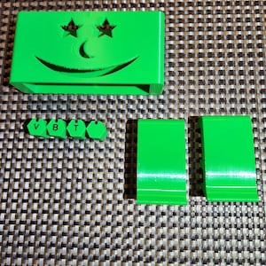 May include: A bright green rectangular object featuring a cutout smiley face with star-shaped eyes and a curved mouth. Included are four small hexagonal pieces with the letters V, B, and T printed on them, alongside two matching green angled stands.
