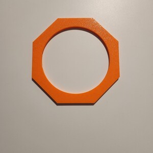May include: A bright orange, flat, octagonal frame with a circular cutout in the center. The object has a textured, matte surface finish and eight straight outer edges, creating a geometric shape that is uniform in color and thickness throughout.