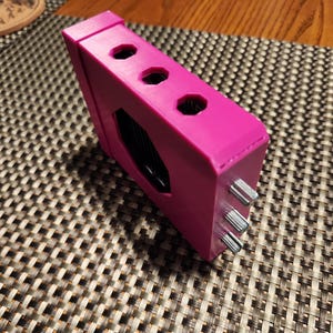 May include: A bright pink rectangular electronic device featuring three circular ports along the top edge and three metallic silver knobs protruding from the side panel. The device has a central black recessed area and a smooth, matte plastic finish.