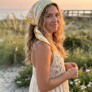 May include: A triangular crochet headscarf featuring alternating horizontal stripes of white and yellow yarn. The headscarf has a delicate scalloped edge detail along the border and is tied at the back with two long, pointed ends.