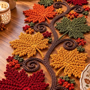 Puede incluir: Un camino de mesa de ganchillo con una enredadera central marrón con zarcillos en espiral, decorado con hojas de arce texturizadas en tonos rojo, naranja, amarillo y verde, acentuado con pequeños racimos de bayas en colores otoñales.