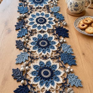 Pode incluir: Um caminho de mesa retangular de crochê com um padrão repetitivo de quatro grandes motivos florais de várias camadas em tons de azul-marinho, azul médio e branco, acentuado com folhas de carvalho em crochê e pequenos elementos brancos.