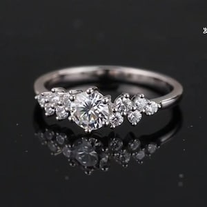 May include: A silver-coloured ring featuring a large, round, clear centre stone flanked by a cluster of smaller, round, clear stones on both sides. The band is smooth and polished, showcasing a reflective surface with a clear, symmetrical design.