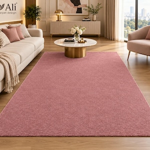 May include: A rectangular area rug in a solid dusty rose pink color with a dense, short-pile texture. The rug features a clean, finished edge and is positioned flat on a light-toned wooden floor, covering a large portion of the visible floor space.