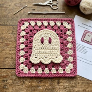 May include: A square crochet granny square featuring a cream-colored ghost motif with dark purple eyes centered on a magenta and cream-colored border. The piece is displayed alongside a metal crochet hook, scissors, and a printed pattern sheet.