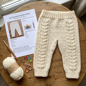 Op de afbeelding: Off-white gehaakte babypantalon met een verticaal kabelpatroon op elke broekspijp, een geribbelde tailleband en geribbelde boorden. Inbegrepen zijn een geprint patroonblad genaamd Leo Snowdrop Baby Pants, een bol zachte wol, een houten haaknaald, een schaar en steekmarkeerders.