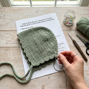 May include: A sage green crocheted baby bonnet with a scalloped edge and long braided ties, resting on a printed pattern titled Classic Baby Bonnet: A Beginner-Friendly Step-by-Step Crochet Pattern (Size 0-6 Months) with yarn, a hook, and scissors.