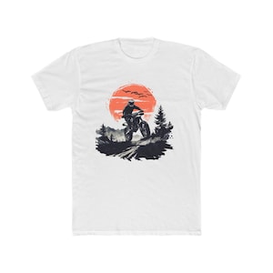 May include: A white short-sleeve t-shirt featuring a graphic print of a motorcyclist riding on a dirt path. The design includes a large orange sun, silhouettes of pine trees, mountains, and birds against a dark, textured ground landscape.