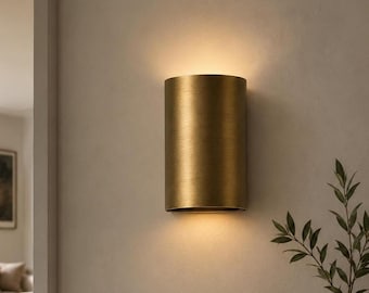 May include: A cylindrical wall sconce with a brushed brass finish. The fixture is mounted vertically and emits a warm, soft glow from both the top and bottom openings, casting light onto the wall surface above and below the metal cylinder.