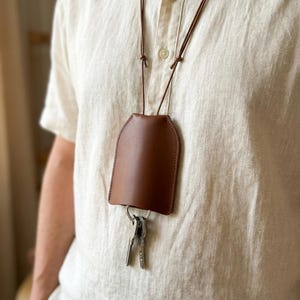 May include: A brown leather key holder pouch suspended from a matching thin leather cord worn around the neck. The smooth, curved pouch features a silver metal split ring at the bottom, which holds two silver-colored metal keys hanging vertically.