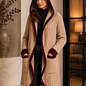 Könnte beinhalten: Eine langärmelige, beige Strickjacke mit Kapuze und Rippenmuster, die einen dunkelroten Besatz an der Kapuze, der Vorderöffnung und den Ärmelbündchen aufweist. Das Kleidungsstück hat zwei große aufgesetzte Taschen mit passenden weinroten Rippkanten.