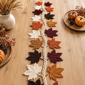 May include: A long, decorative table runner featuring a central cream-colored crocheted vine with attached crocheted maple leaves in various autumn shades including deep burgundy, burnt orange, golden yellow, dark brown, and off-white.
