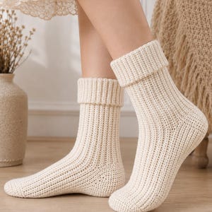 May include: A pair of cream-colored, mid-calf length socks featuring a thick, vertical ribbed knit texture and a wide, folded cuff at the top. The socks are constructed with a dense, uniform stitch pattern that covers the entire foot and ankle area.