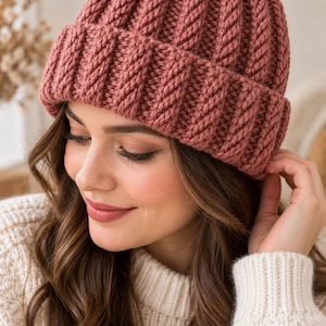 May include: A dusty rose beanie crafted with a thick, vertical cable knit pattern. The hat features a wide, folded cuff that encircles the base, providing a textured, ribbed appearance throughout the entire construction of the headwear piece.