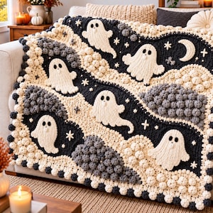 May include: A crochet blanket featuring a repeating pattern of white ghosts, dark gray textured clouds, and small white stars on a black background. The blanket has a thick, bumpy border made of alternating cream and dark gray crochet spheres.