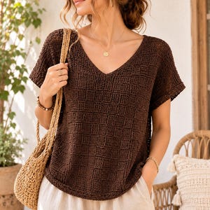 May include: A dark brown short-sleeved knit top featuring a textured checkerboard pattern and a V-neckline, paired with light beige trousers and a woven straw-colored tote bag with braided handles, worn with a gold necklace and gold hoop earrings.