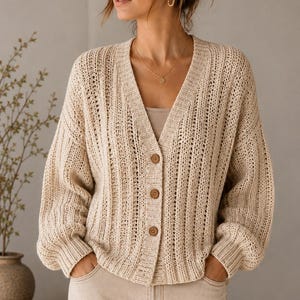 May include: A beige, long-sleeved, open-knit cardigan featuring a V-neckline and three brown buttons down the front. The garment has a ribbed texture, dropped shoulders, and a relaxed fit, worn over a simple beige top and light-coloured trousers.