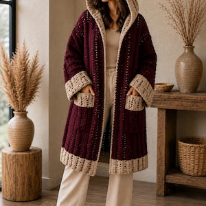 May include: A long-sleeved, knee-length crochet cardigan in deep burgundy with a contrasting beige hood, cuffs, hem, and two front pockets. The garment features a vertical ribbed texture throughout the main body and an open front design.