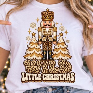 Può includere: Una t-shirt bianca a maniche corte con la grafica di uno schiaccianoci tra due alberi di Natale, tutto con una stampa leopardata marrone. Il testo sottostante recita WILD LITTLE CHRISTMAS in un font retrò marrone con accenti a stella dorati.