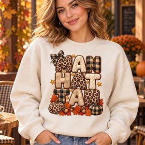 Könnte beinhalten: Ein cremefarbenes Sweatshirt mit Rundhalsausschnitt und dem Wort MANHATTAN in großen Blockbuchstaben. Jeder Buchstabe hat ein einzigartiges Muster, darunter Leopardenmuster, Karomuster und Zopfstrick, verziert mit kleinen Kürbissen und Blättern.
