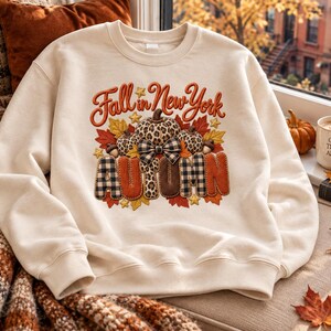 Könnte beinhalten: Ein cremefarbenes Sweatshirt mit Rundhalsausschnitt und einem Grafikdesign mit dem Text Fall in New York und AUTUMN. Das Motiv zeigt Kürbisse mit Leopardenmuster und Karomuster, eine karierte Schleife, Eicheln sowie orange und gelbe Herbstblätter.