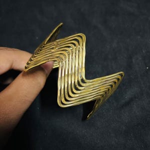 May include: A gold-toned metal cuff bracelet featuring a repeating zigzag or chevron pattern. The design consists of multiple parallel, thin, wavy metal wires that create a sharp, angular silhouette with a textured, reflective surface finish.