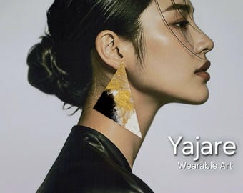 Può includere: Un grande orecchino triangolare caratterizzato da un design astratto con foglia d'oro testurizzata, vernice nera e bianca. L'orecchino è indossato su un orecchio e il testo "Yajare Wearable Art" è visibile nell'angolo in basso a destra.