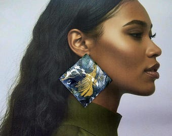 Peut inclure: Une grande boucle d'oreille de forme carrée présentant un motif marbré abstrait dans des tons bleu marine profond, blanc et or métallique. La surface a une finition brillante avec des textures tourbillonnantes visibles et distinctes.
