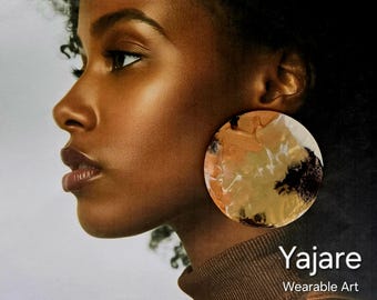 Può includere: Un grande orecchino circolare con una superficie astratta multicolore caratterizzata da vortici color marrone chiaro, crema e nero. L'orecchino è indossato sul lobo. Il testo "Yajare Wearable Art" è stampato in bianco nell'angolo in basso a destra.
