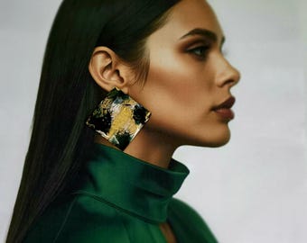 Peut inclure: Une grande boucle d'oreille carrée présentant un motif peint abstrait avec des traits audacieux de noir, d'or métallique et de blanc. La boucle d'oreille a une forme géométrique plate et une finition de surface texturée, fixée au lobe.