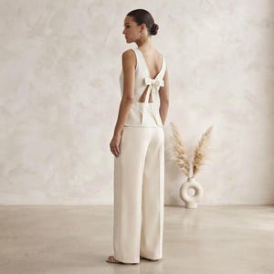 May include: A cream-colored sleeveless top featuring a deep V-back with a central bow detail, paired with matching wide-leg trousers. The top has a fitted bodice that transitions into a clean, straight silhouette, while the trousers fall straight.