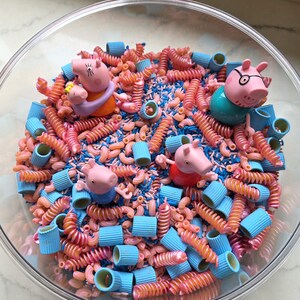 May include: A clear glass bowl filled with a sensory bin mixture of pink and blue dyed pasta shapes, including spiral, macaroni, and tube noodles, interspersed with four small pink pig figurines wearing colourful clothing and glasses.