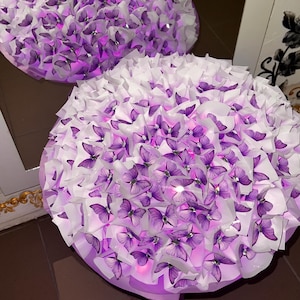 Op de afbeelding: Een rond boeket met talloze paarse vlindervormige ornamenten afgewisseld met witte papieren lagen. Het arrangement wordt van binnenuit verlicht door kleine lampjes, wat een gloeiend effect creëert door de gehele paarse en witte structuur.