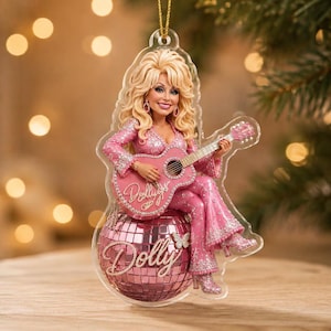Könnte beinhalten: Ein hängendes Ornament mit einer blonden Figur in einem glitzernden rosa Outfit, die eine rosa Akustikgitarre spielt. Die Figur sitzt auf einer rosa Discokugel, auf der der Name Dolly in Schreibschrift steht, ebenso wie auf der Gitarre.