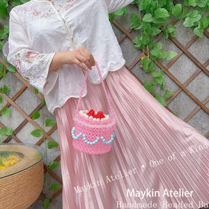 May include: A cylindrical beaded handbag shaped like a cake, featuring a pink body, a light blue beaded scalloped trim, and red and white beaded berries on top. The bag has a single beaded handle and is held against a white floral blouse and pink skirt.