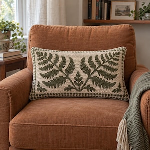 May include: A rectangular crochet pillow featuring a cream-colored background with a dark green fern leaf pattern. The design includes a symmetrical arrangement of two large fern fronds and a smaller central sprig, bordered by a textured edge.