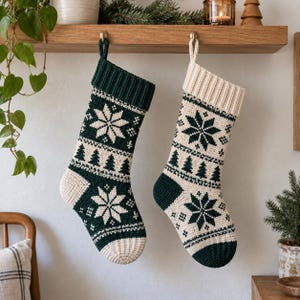 Könnte beinhalten: Zwei gehäkelte Weihnachtsstrümpfe hängen nebeneinander. Der linke Strumpf ist dunkelgrün mit cremefarbenen Schneeflocken- und Tannenbaummustern. Der rechte Strumpf ist cremefarben mit dunkelgrünen Schneeflocken- und Tannenbaummustern.
