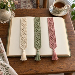 May include: Three rectangular crochet bookmarks laid flat on an open book. Each bookmark features a delicate leaf-like lace pattern and a matching tassel at the bottom. The bookmarks are coloured in cream, sage green, and dusty rose, respectively.