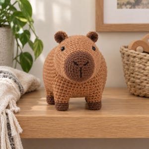 May include: A crochet capybara plush toy featuring a light brown body, a darker brown snout, and small dark brown feet. The stuffed animal has black bead eyes and a stitched nose and mouth detail, crafted with a textured yarn stitch pattern.