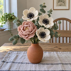 May include: A bouquet of crocheted flowers in a terracotta vase. The arrangement features one large dusty pink peony, three white anemones with dark centers, and several green crocheted eucalyptus stems with round leaves, all displayed in a simple pot.