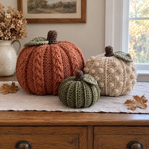 May include: Three handmade crochet pumpkins in varying sizes and colors. The largest is burnt orange with a cable knit texture, the medium is cream with a bobble stitch pattern, and the smallest is olive green with vertical ribbed stitching.
