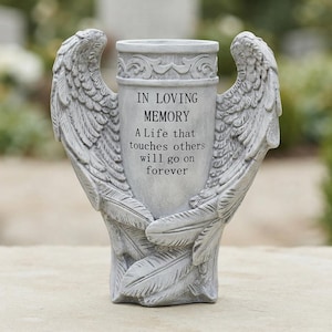 Op de afbeelding: Een grijze gedenkvaas in steenlook met aan beide zijden gebeeldhouwde engelvleugels. De voorzijde is gegraveerd met de tekst: IN LOVING MEMORY, A Life that touches others will go on forever. De cilindrische vaas heeft een decoratieve bovenrand.