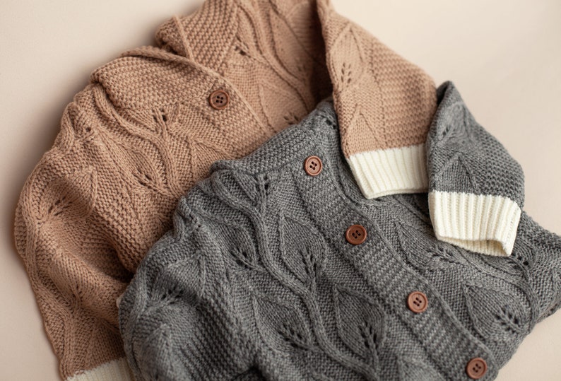 May include: Two knitted cardigans with leaf patterns. One is brown and the other is gray. Both have white trim on the cuffs and collar.