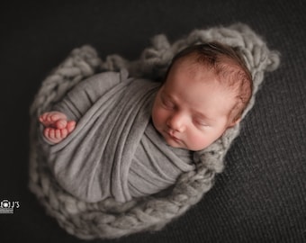 May include: A newborn baby is swaddled in a gray blanket and sleeping peacefully. The baby is lying on a gray knitted surface.