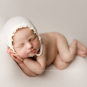 May include: A newborn baby wearing a white crocheted bonnet sleeps on a white background. The baby is lying on its side with its head resting on its arms.